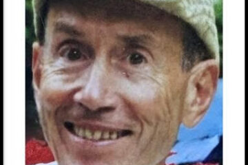 looking directly into the camera and smiling while sporting a traveler's cap