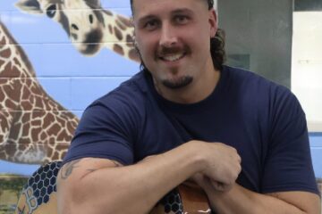 fella holding his guitar and smiling at the camera. a painting of a giraffe looking at us over his shoulder