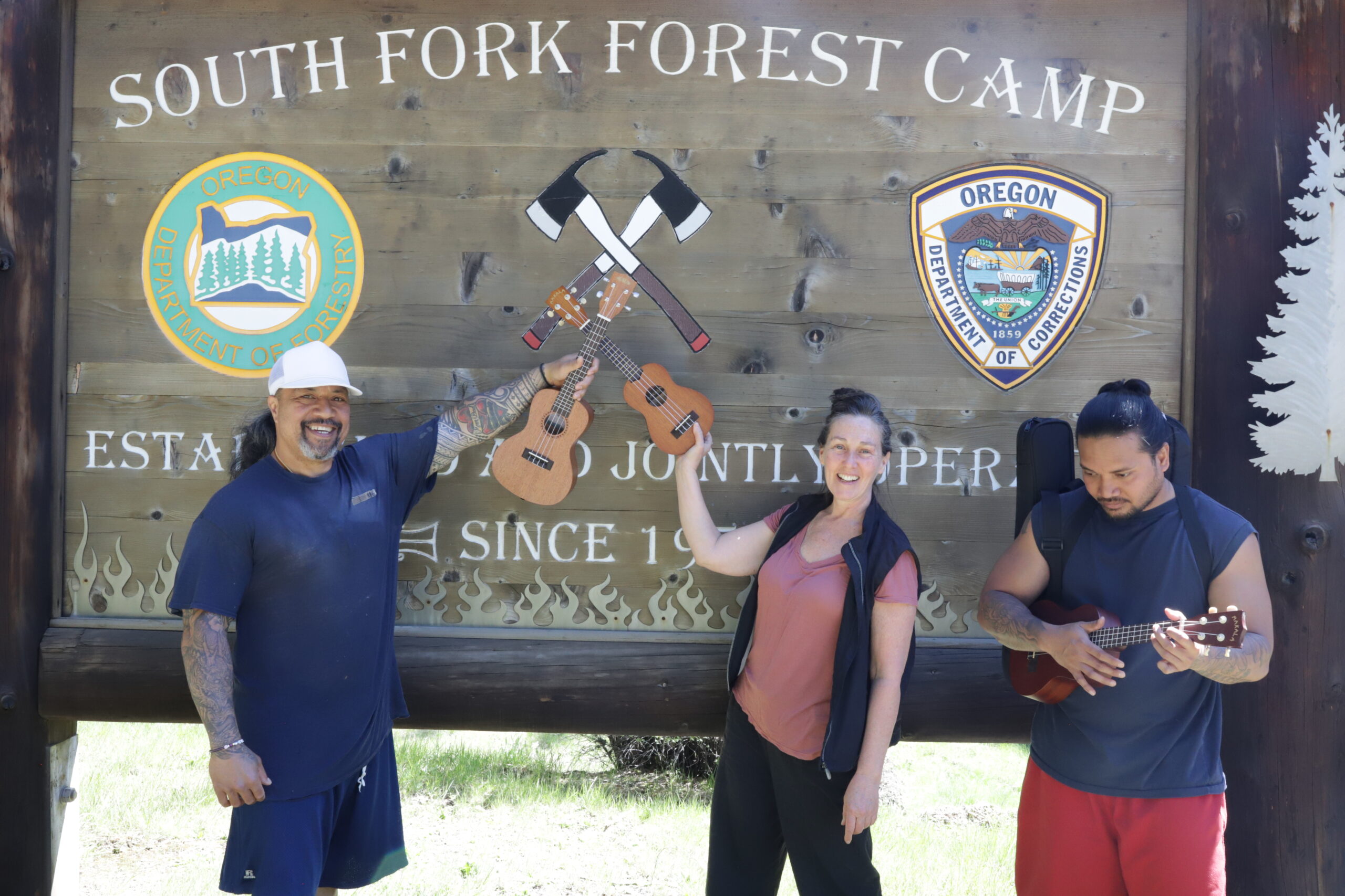 A smiling man and woman hold up ukuleles in front of a wood sign that reads "South Fork Forest Camp" while another man concentrates on playing a ukulele beside them