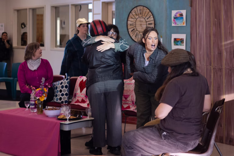 scene of 6 women in a living room, 2 hug, 1 makes a face at another behind their back, 1 looks to another expectanly
