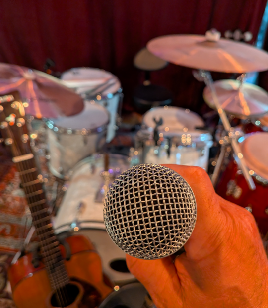 Mans hand holding a microphone in front of a drum set.