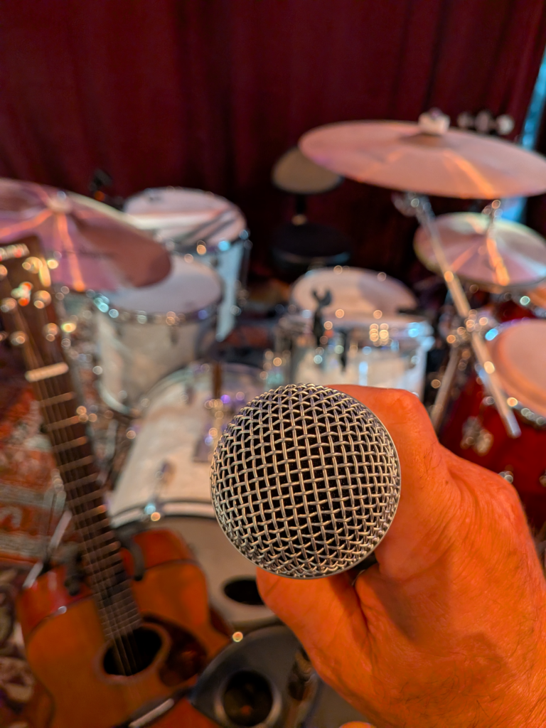 Mans hand holding a microphone in front of a drum set.