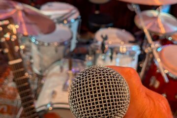 Mans hand holding a microphone in front of a drum set.